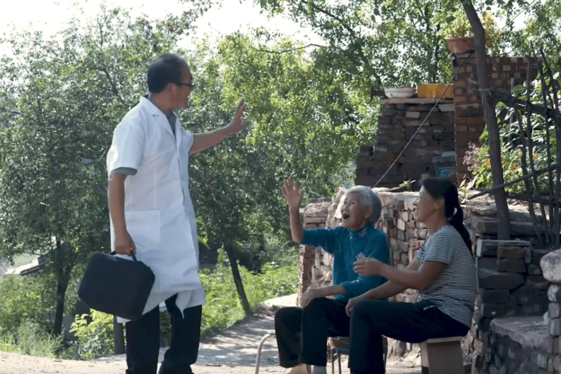 A male doctor walking along a path waves to two women sitting on a bench. The women smile and wave back.