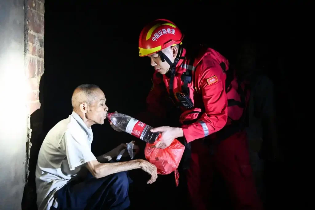 A rescue worker stands in front of an elderly man holding a bottle of water for the man to drink